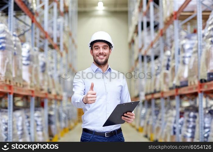 wholesale, logistic, business, export and people concept - happy man in hardhat with clipboard at warehouse showing thumbs up gesture