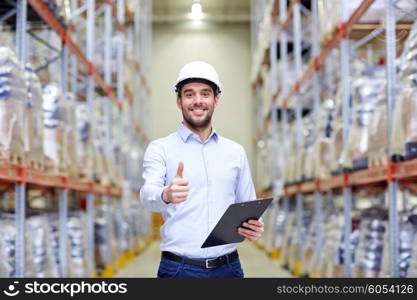 wholesale, logistic, business, export and people concept - happy man in hardhat with clipboard at warehouse showing thumbs up gesture