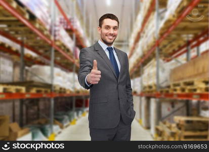 wholesale, logistic, business, export and people concept - happy man in suit and tie showing thumbs up gesture over warehouse background