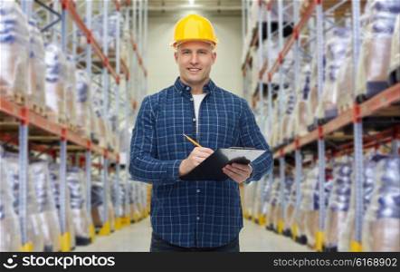 wholesale, logistic, business, export and people concept - happy man or manager in helmet with clipboard and pencil checking goods over warehouse background