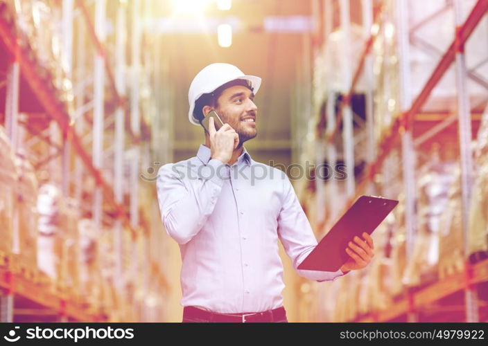 wholesale, logistic, business, export and people concept - smiling businessman with clipboard calling on smartphone at warehouse. man with clipboard and smartphone at warehouse