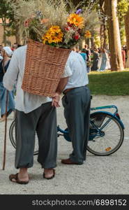 Wicker Basket Filled with Sheafs of wheat and Flowers Carried over the Shoulder of Countryman and Old Man with Bicycle