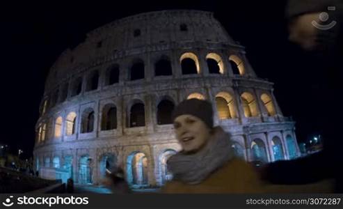 Wide angle shot of loving tourist couple coming up to Coliseum at night, making selfie with cell phone and leaving