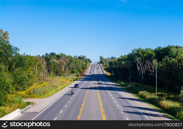 Wide multi-lane road with few cars in wooded area on blue sky.