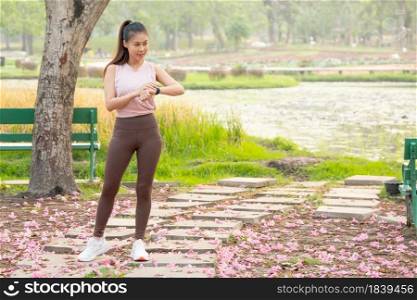 Wide shot of Asian woman stand and look at her watch during exercise in park or garden with green tree and lake as background in the morning.