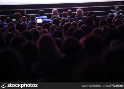 Wide shot of sihouettes of people from back watching cinema or performance. Somebody filming the show using tablet