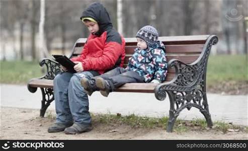 Wide shot of young boy watching his brother as the two sit together on a wooden park bench in warm winter clothing with the older child using a tablet computer