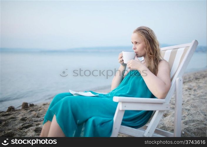 Wide shot of young girl enjoying a cup of tea at the seaside sitting relaxing on a deckchair with a blissful expression overlooking a tropical beach