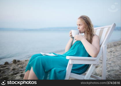 Wide shot of young girl enjoying a cup of tea at the seaside sitting relaxing on a deckchair with a blissful expression overlooking a tropical beach