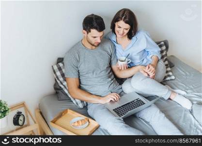 Wife and husband make shopping online, choose present for their little child in internet, look at laptop computer, enjoy togetherness in bedroom, drink hot beverage. People, leisure, technology