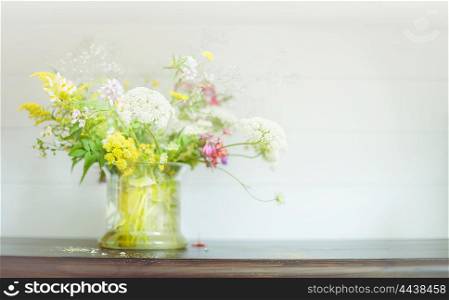 Wild flowers bunch in glass pot on wooden shelf at light background. Floral Home decoration and interior