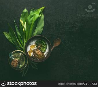 Wild garlic yogurt dip or dressing in wooden bowl on dark rustic kitchen table background with ingredients, top view with copy space. Healthy seasonal food concept