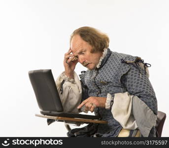 William Shakespeare in period clothing sitting in school desk with laptop computer and hand to head looking perplexed.