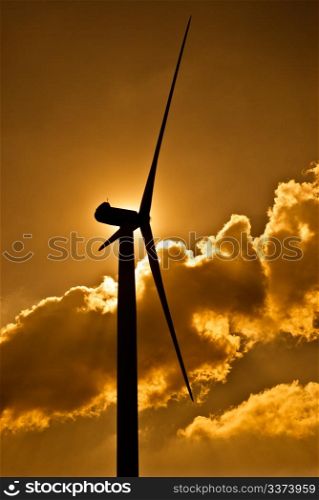 Wind turbine in the back light of the sun with yellow sky and clouds.