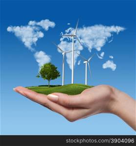 Wind turbines on meadow with tree holds in womans hand against blue sky and map of the world made of clouds. Worldwide Green energy concept. Wind turbines with tree in female hand