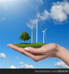 Wind turbines on meadow with tree holds in womans hand against blue sky and clouds. Green energy concept. Wind turbines with tree in female hand