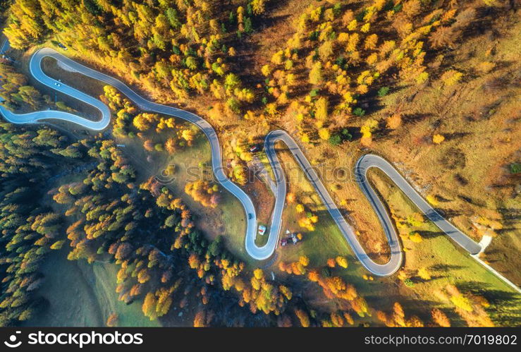 Winding road in autumn forest at sunset in mountains. Aerial view. Top view of beautiful asphalt roadway and orange trees. Highway through the woodland in fall. Trip in europe. Travel and nature