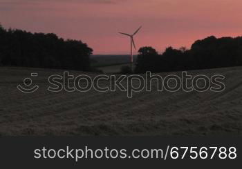 Windrad dreht sich im Abendrot, davor ein geerntetes Getreidefeld und die Schatten der BSume