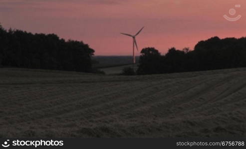 Windrad dreht sich im Abendrot, davor ein geerntetes Getreidefeld und die Schatten der BSume