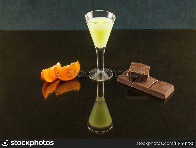 Wineglass on a thin stalk with a drink, chocolate and tangerine on a mirror surface with reflection