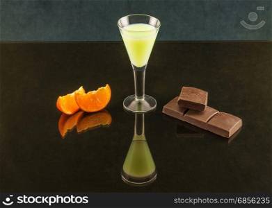 Wineglass on a thin stalk with a drink, chocolate and tangerine on a mirror surface with reflection