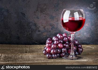 Wineglass with red wine and red grapes on wooden table. Dark background.. Wineglass with red wine