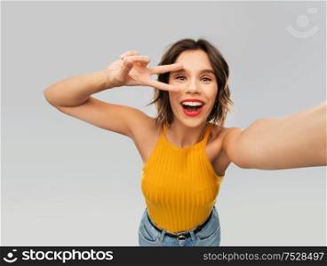 winning gesture, emotions and people concept - happy smiling young woman in mustard yellow top taking selfie and showing peace hand sign over grey background. happy woman taking selfie and showing peace sign