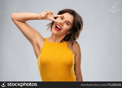 winning gesture, emotions and people concept - happy smiling young woman in mustard yellow top showing peace hand sign over grey background. happy smiling young woman showing peace hand sign