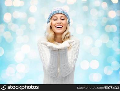winter, advertisement, christmas and people concept - smiling young woman in winter hat and sweater holding something on her empty palms over blue holidays lights background