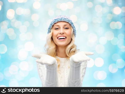 winter, advertisement, christmas and people concept - smiling young woman in winter hat and sweater holding something on her empty palms over blue holidays lights background