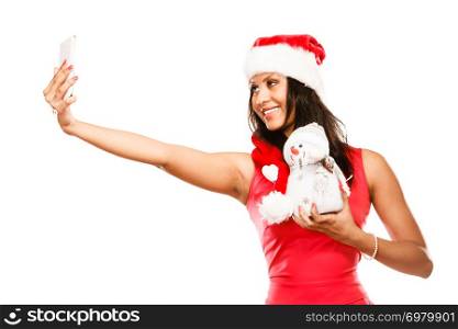 Winter and Christmas time concept. Woman in santa helper hat, mixed race girl in red dress holding happy nice snowman toy, taking self picture selfie with smartphone camera. Studio shot on white