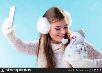 Winter and Christmas time concept. Woman teen girl in warm clothing holding happy nice snowman toy, taking self picture selfie with smartphone camera. Studio shot on blue background