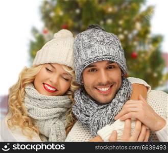 winter and holidays concept - happy couple in hats and scarves hugging over christmas tree background. happy couple hugging over christmas tree