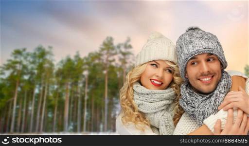 winter, christmas and people concept - happy couple in hats and scarves hugging over forest background. couple hugging over winter forest background
