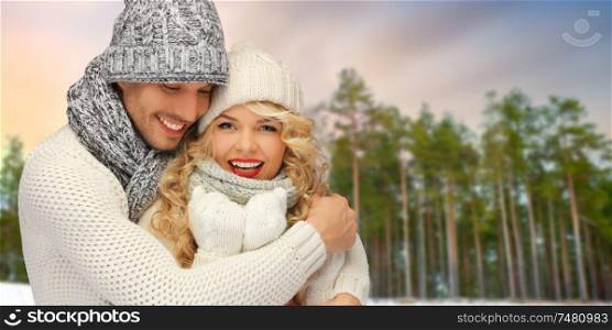 winter, christmas and people concept - happy couple in hats and scarves hugging over forest background. couple hugging over winter forest background