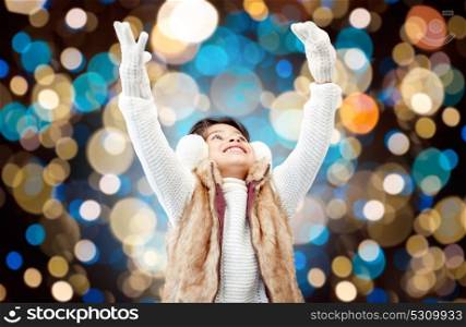 winter, christmas and people concept - happy little girl wearing earmuffs and gloves over holidays lights background. happy girl in winter earmuffs over holidays lights