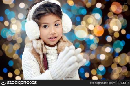 winter, christmas and people concept - happy little girl wearing earmuffs and gloves over holidays lights background. girl in winter earmuffs over holidays lights