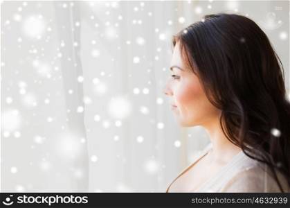 winter, christmas, people and leisure concept - young woman looking through window at home over snow