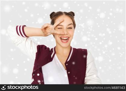 winter, christmas, people, gesture and teens concept - happy smiling pretty teenage girl showing peace sign and winking over gray background and snow