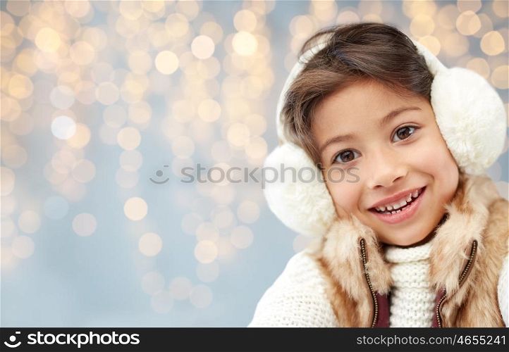winter, christmas, people, happiness concept - happy little girl wearing earmuffs over holidays lights background