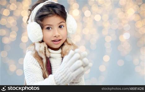 winter, christmas, people, happiness concept - happy little girl wearing earmuffs over holidays lights background