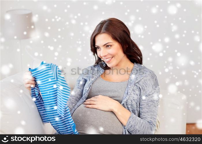winter, christmas , pregnancy and people concept - happy woman holding and looking at blue baby boys bodysuit at home over snow