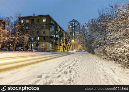 Winter cityscape with a snow-covered road along a city street with residential buildings and trees in the snow, with city evening illumination against the backdrop of blue twilight.. Snow-covered road along a city street with trees in the snow and city evening lighting against the backdrop of blue twilight.