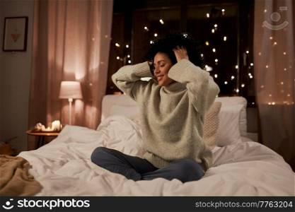 winter, comfort and people concept - happy smiling woman in woolen high neck sweater touching her hair in bed at home at night. woman in sweater touching her hair in bed at night