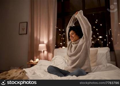 winter, comfort and people concept - happy smiling woman in woolen high neck sweater stretching in bed at home at night. woman in sweater stretching in bed at night