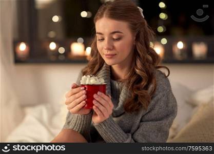 winter, comfort and people concept - happy young woman in pullover holding red mug with whipped cream in bed at night. happy woman holding mug with whipped cream in bed