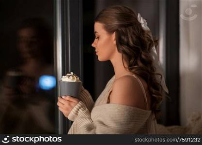winter, comfort and people concept - young woman in pullover holding mug with whipped cream at window at night. woman holding mug with whipped cream at night