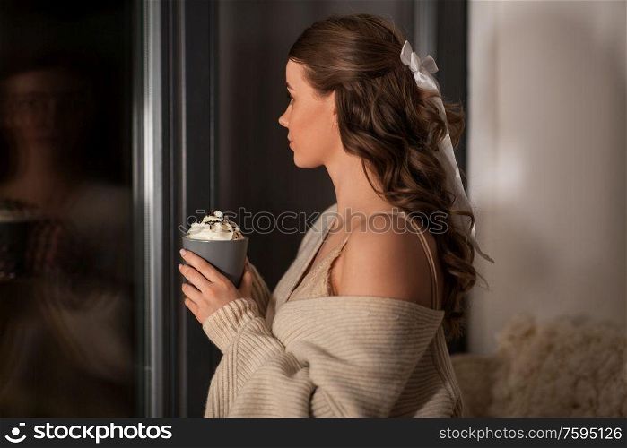 winter, comfort and people concept - young woman in pullover holding mug with whipped cream at window at night. woman holding mug with whipped cream at night