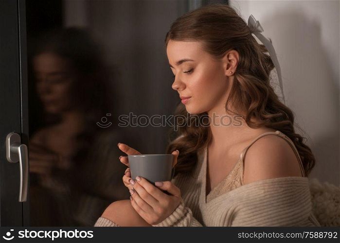 winter, comfort and people concept - young woman in pullover sitting at window with coffee or tea cup at home. woman with coffee or tea cup at window at home