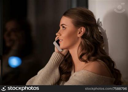 winter, comfort and people concept - young woman in pullover sitting at window at home and calling on smartphone at night. woman calling on smartphone at window at night